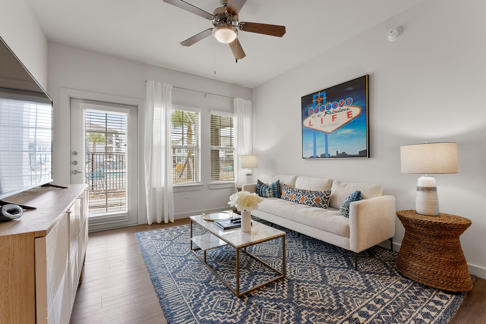 A cozy living room featuring a white sofa with decorative pillows, a coffee table, a woven side table, and a colorful wall art piece. Large windows allow natural light in, with a view of an outdoor space.