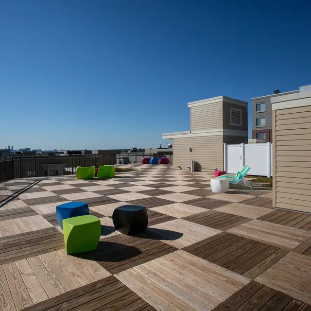 Bright Rooftop Deck with Colorful Seating A rooftop deck with a checkerboard pattern made of wooden planks. Colorful seating options are scattered across the area, including green, blue, pink, and black seating. The skyline can be seen in the distance under a clear blue sky.