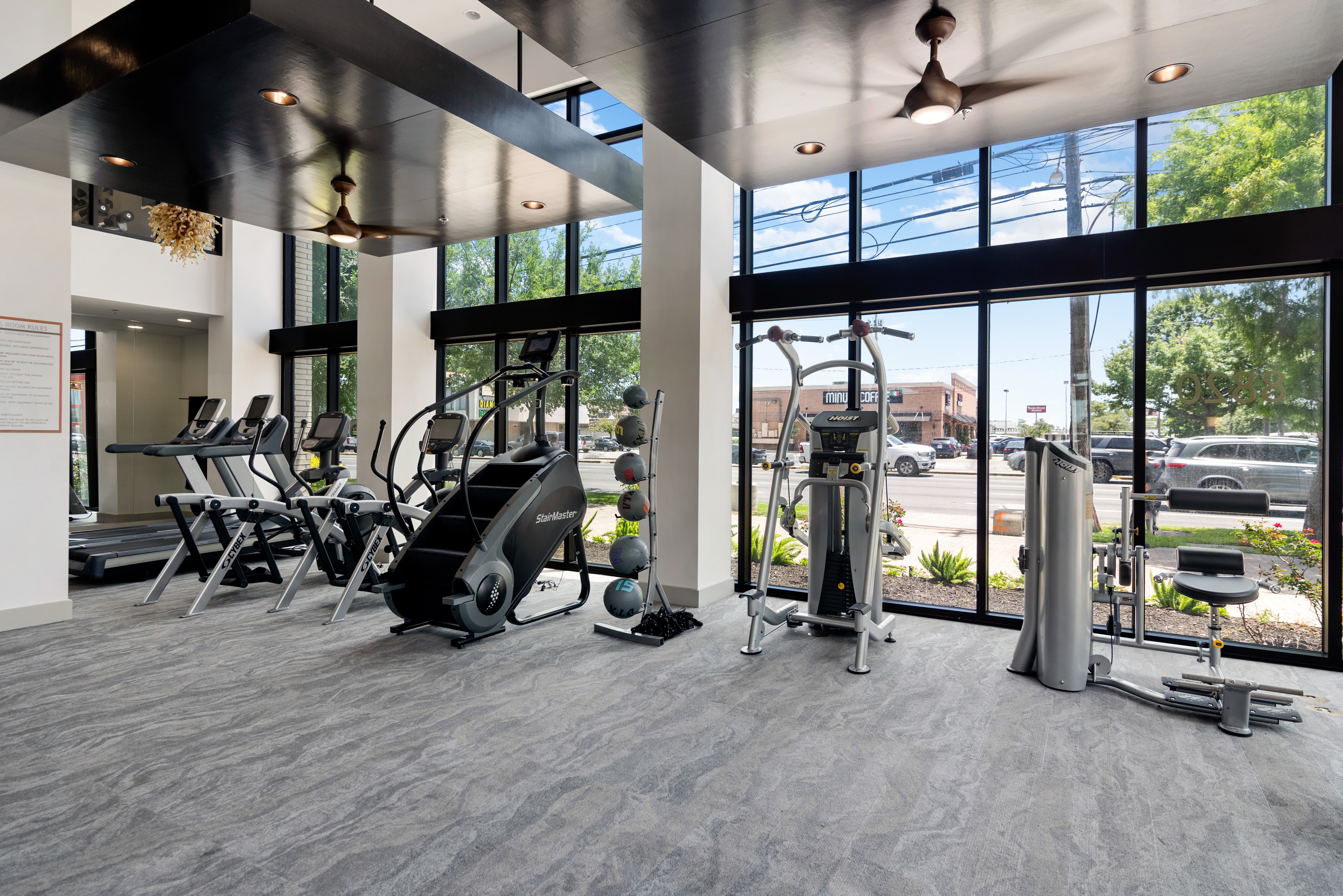A modern gym interior with various fitness equipment including treadmills, ellipticals, and strength training machines, illuminated by natural light from large windows.