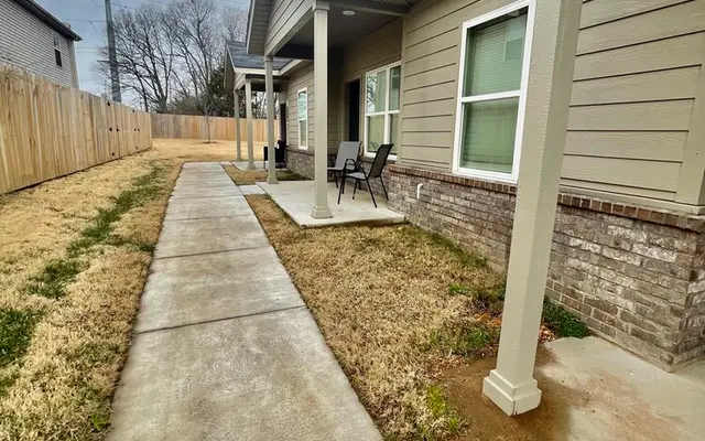 Pathway alongside a residential home with a well-kept lawn and chairs on a porch.