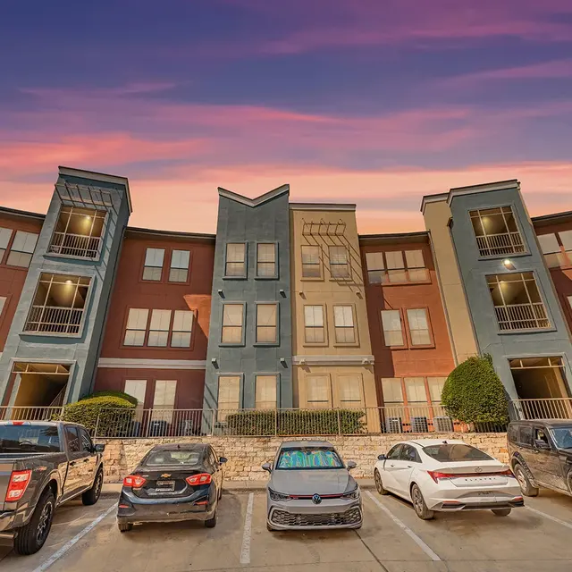 A multi-story apartment complex with balconies and a parking lot in front, captured during sunset with a pink and purple sky.