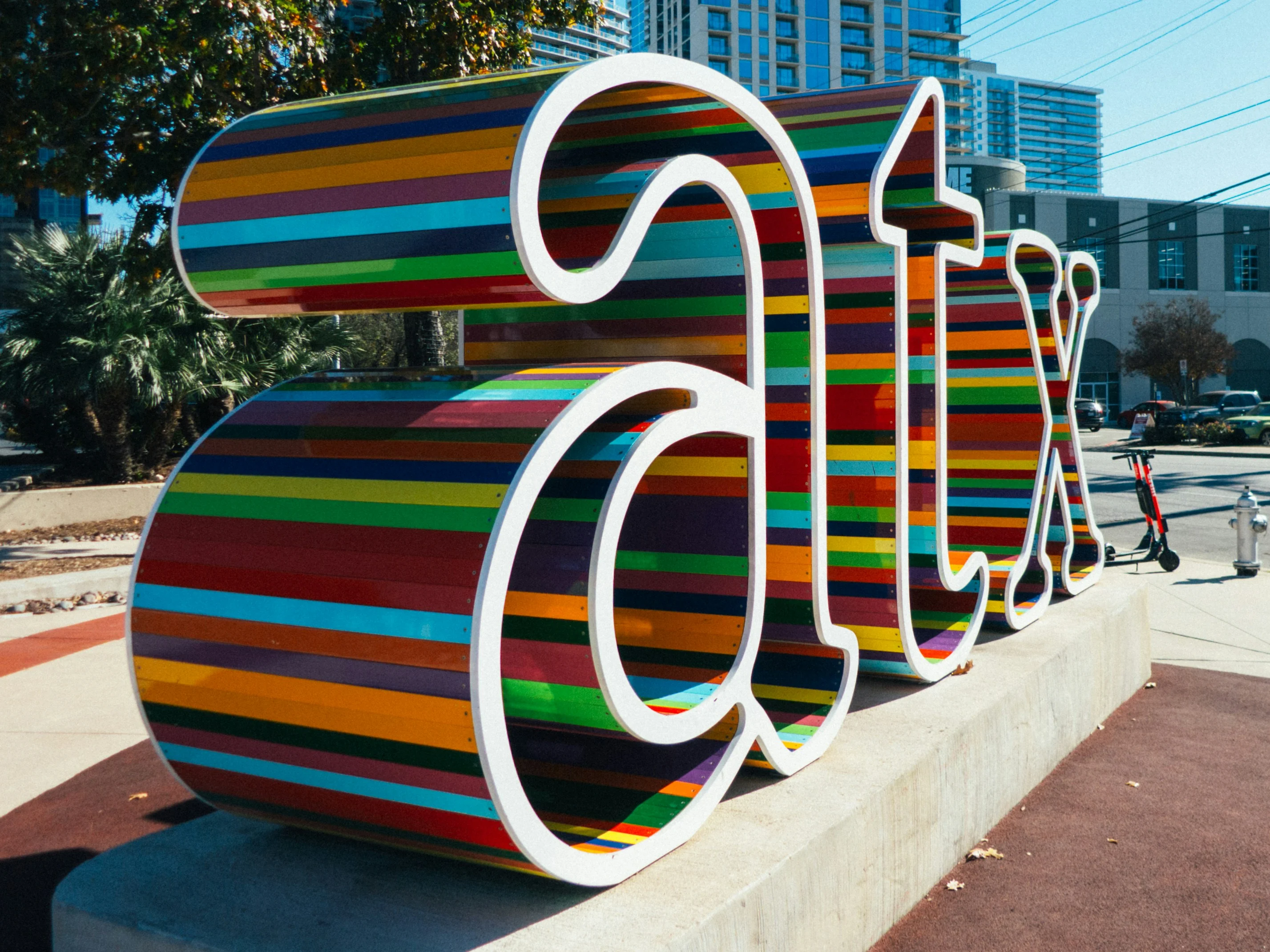 Colorful ATX Sign A large, colorful sign with the letters 'atx' in a playful, bold font. The letters are designed with a striped pattern in various vibrant colors, set against a concrete base, with a background of trees and buildings.