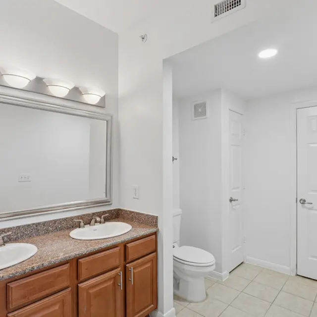 A well-lit modern bathroom featuring a double vanity with two sinks, a large mirror, wooden cabinetry, tiled flooring, and a separate toilet area behind a door.