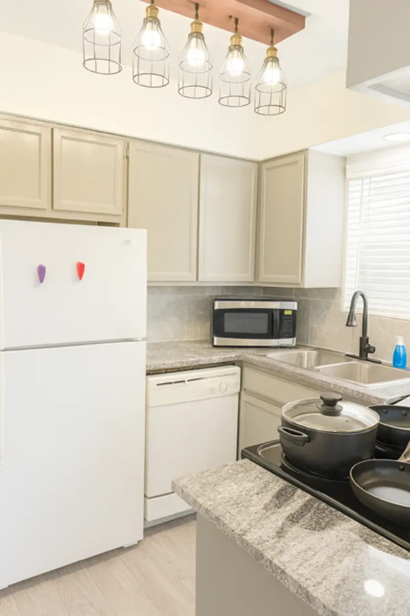 Modern Kitchen Design A modern kitchen with light-colored cabinets, a white refrigerator, a microwave, a sink with a modern faucet, and a black stovetop with cooking pots. The ceiling features hanging light fixtures, and there are windows with blinds.
