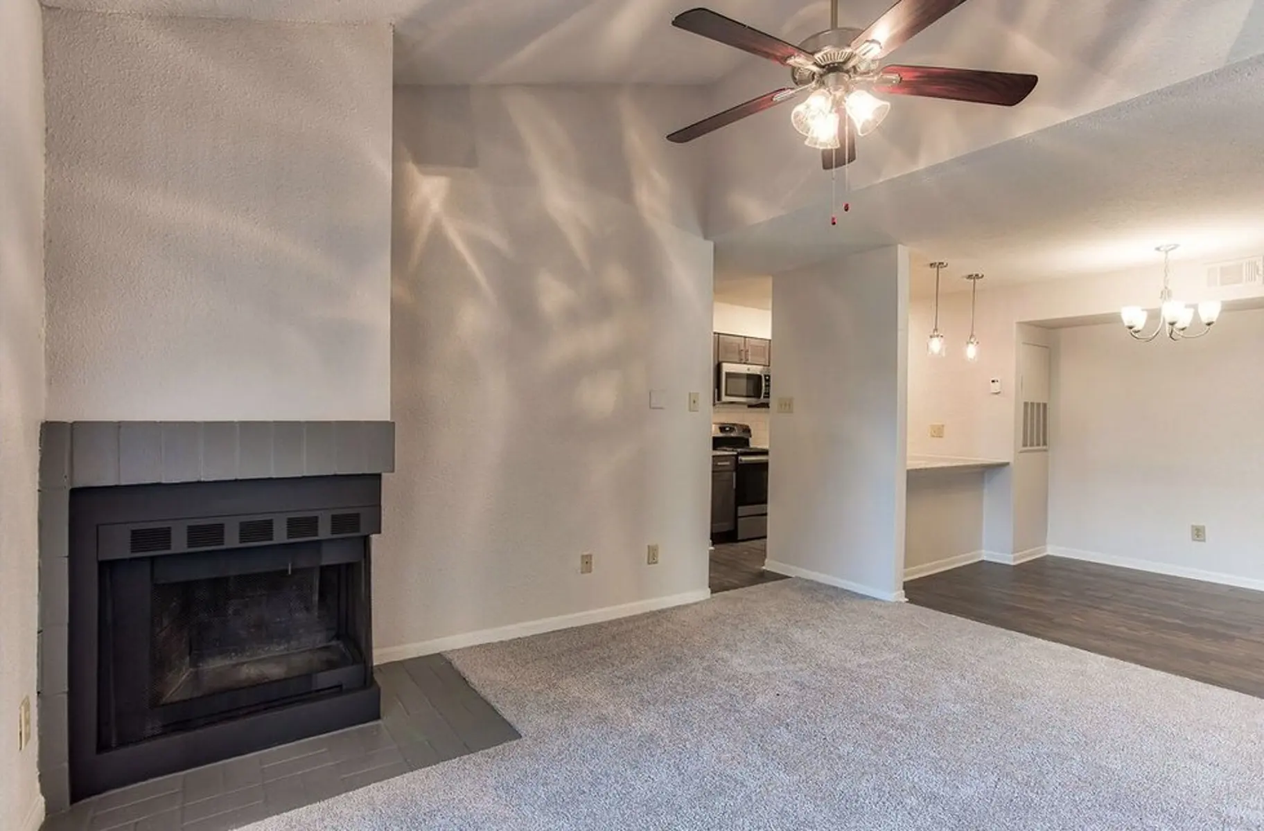 A spacious living room featuring a gray carpeted floor, a fireplace with a black outer frame, a ceiling fan with light fixtures, and a view into a kitchen area with modern appliances. The walls are painted white, and there is a subtle light reflection pattern on the wall.