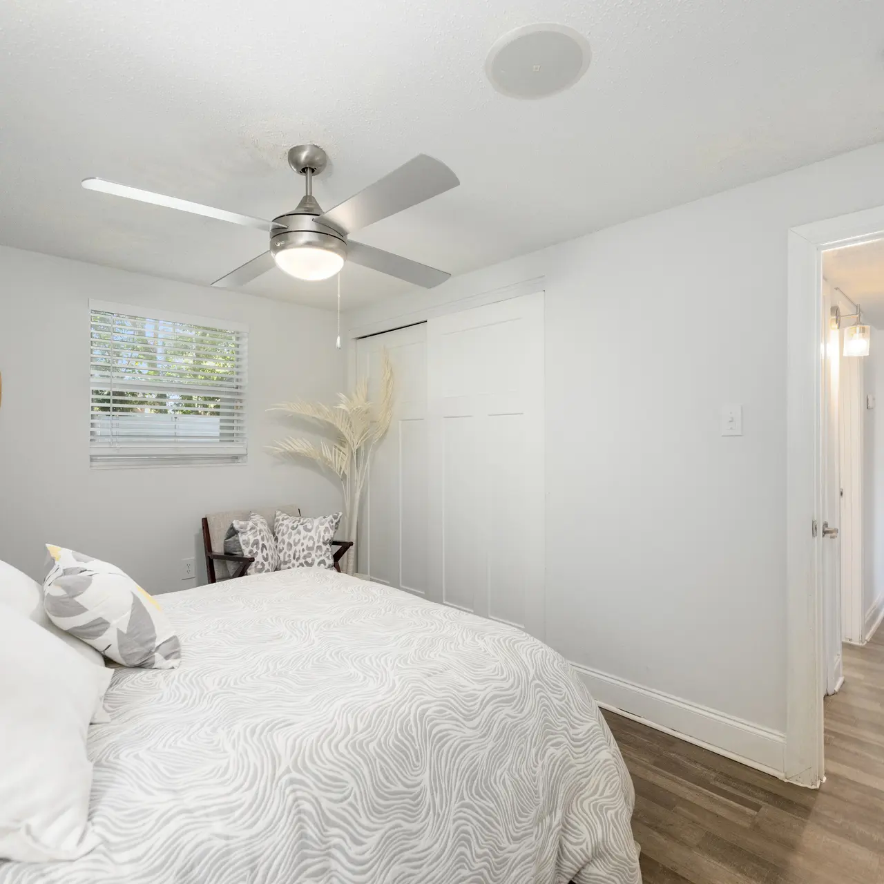 A modern bedroom featuring a bed with light-colored bedding and decorative pillows. There is a ceiling fan, a circular mirror on the wall, and a small chair next to the bed. The floor is wooden, and there's a doorway leading to another room.