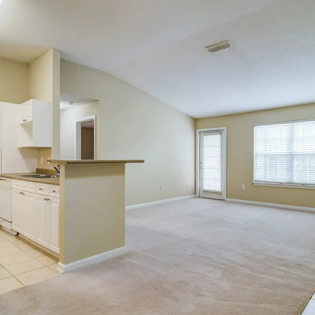 An open concept living area featuring a kitchen and living room with beige walls and carpet. The kitchen has white cabinets and tile flooring, while the living room has large windows allowing natural light. The space is well-lit and airy.