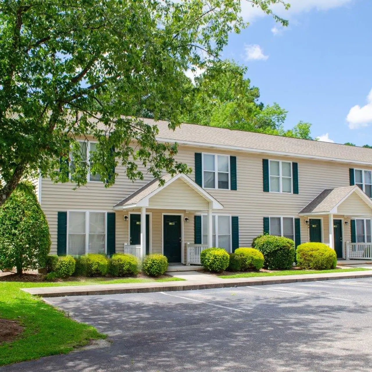 A row of beige two-story townhouses with green shutters and white trim, surrounded by landscaping and trees. The scene is set under a bright blue sky with white clouds.