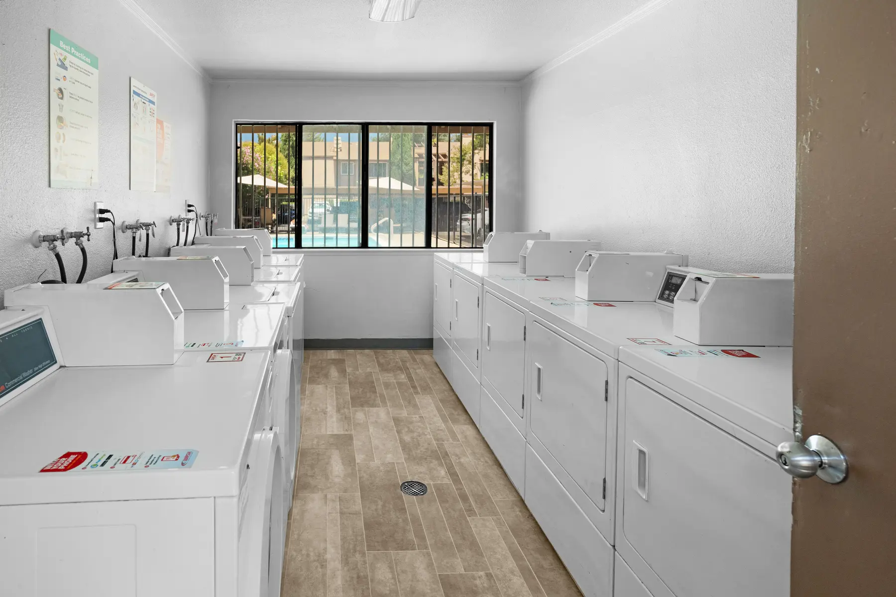 Interior view of a laundry room with multiple washing machines and dryers lined up against the walls, floor covered with wood-like tiles, and a large window overlooking a pool area.