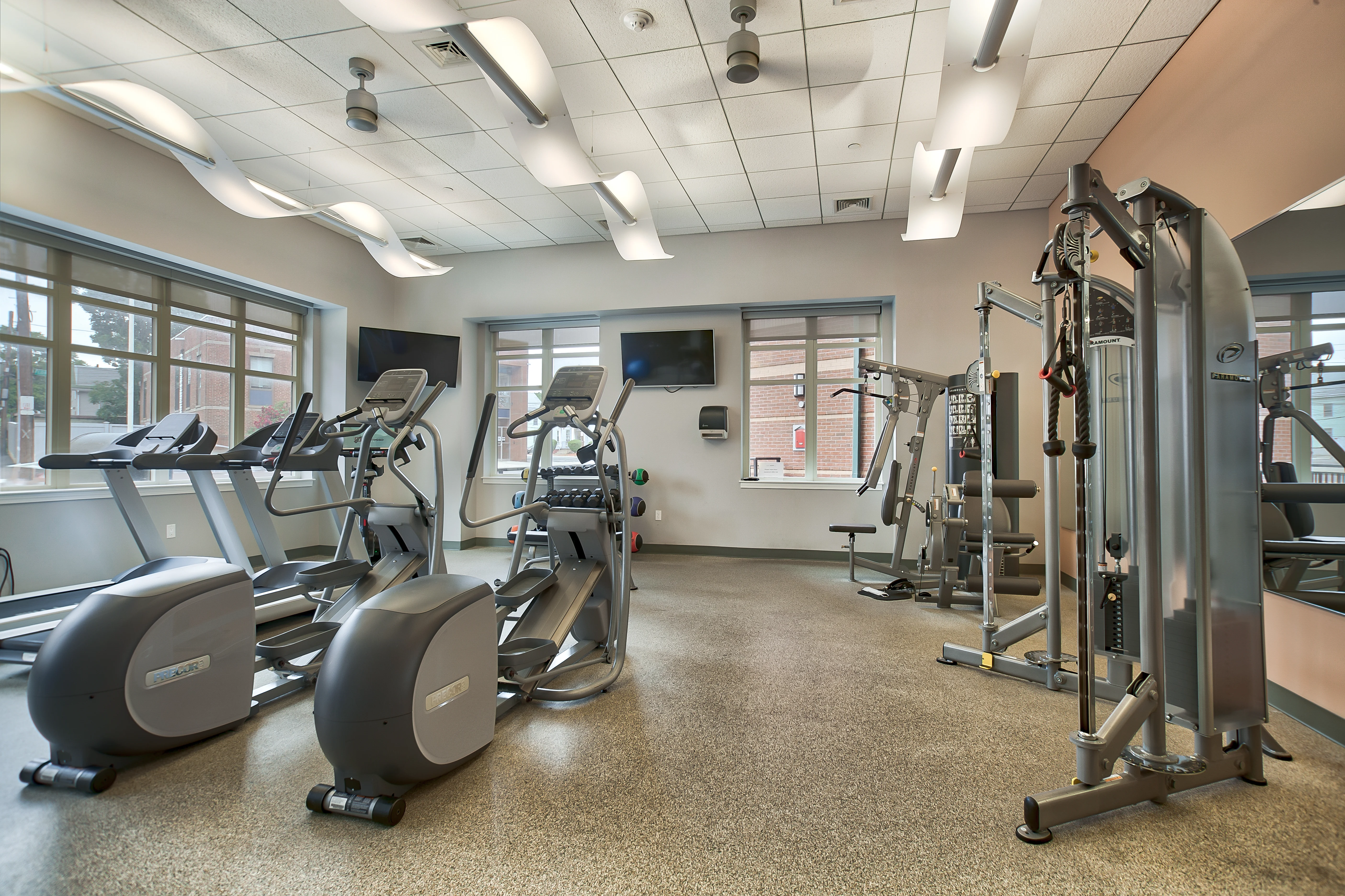 A modern gym interior featuring various exercise equipment including treadmills, ellipticals, and a multi-station weight machine, with large windows allowing natural light.