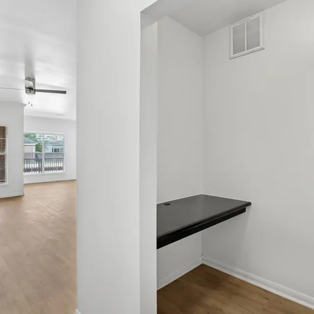 A modern apartment interior with a living area visible through an archway. The living space features large windows, a ceiling fan, and light-colored flooring. To the right, a small room with a black countertop and a closet is partially visible.