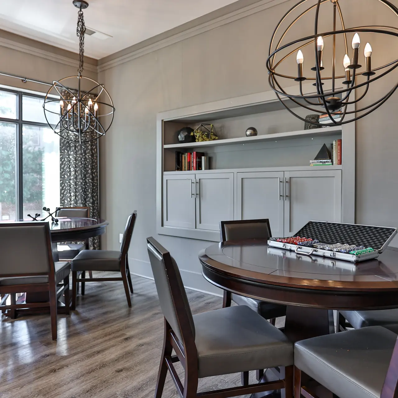Elegant Modern Dining Space A modern dining area featuring two round tables with eight dark wooden chairs, elegant pendant lights hanging from the ceiling, and a wall-mounted bookshelf filled with decorative items. Large windows with patterned curtains allow natural light to enter the space.