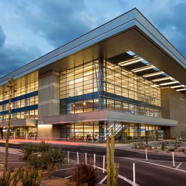 A modern airport terminal building at dusk with large glass windows and contemporary architecture. Palm trees and desert vegetation are in the foreground, and a cloudy sky looms overhead.