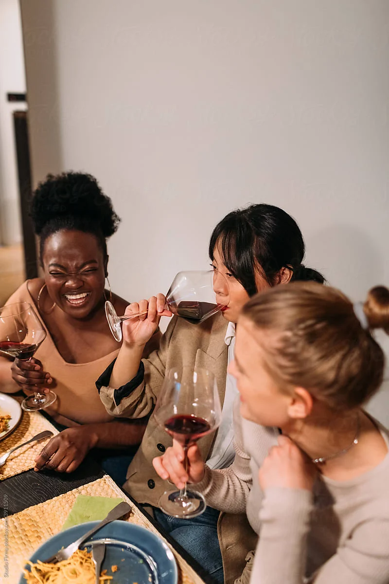 A group of three women enjoying a dinner together, laughing and toasting with glasses of red wine. One woman is playfully sipping from a glass while the others look on joyfully. Plates of food, including pasta, are visible on the table.