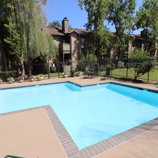 A view of a swimming pool in a landscaped area with green trees and apartment buildings in the background.