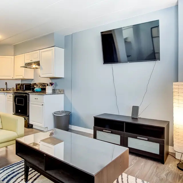 A modern living room with a couch, a coffee table, a television mounted on the wall, and a lamp. There is a kitchen area visible in the background.