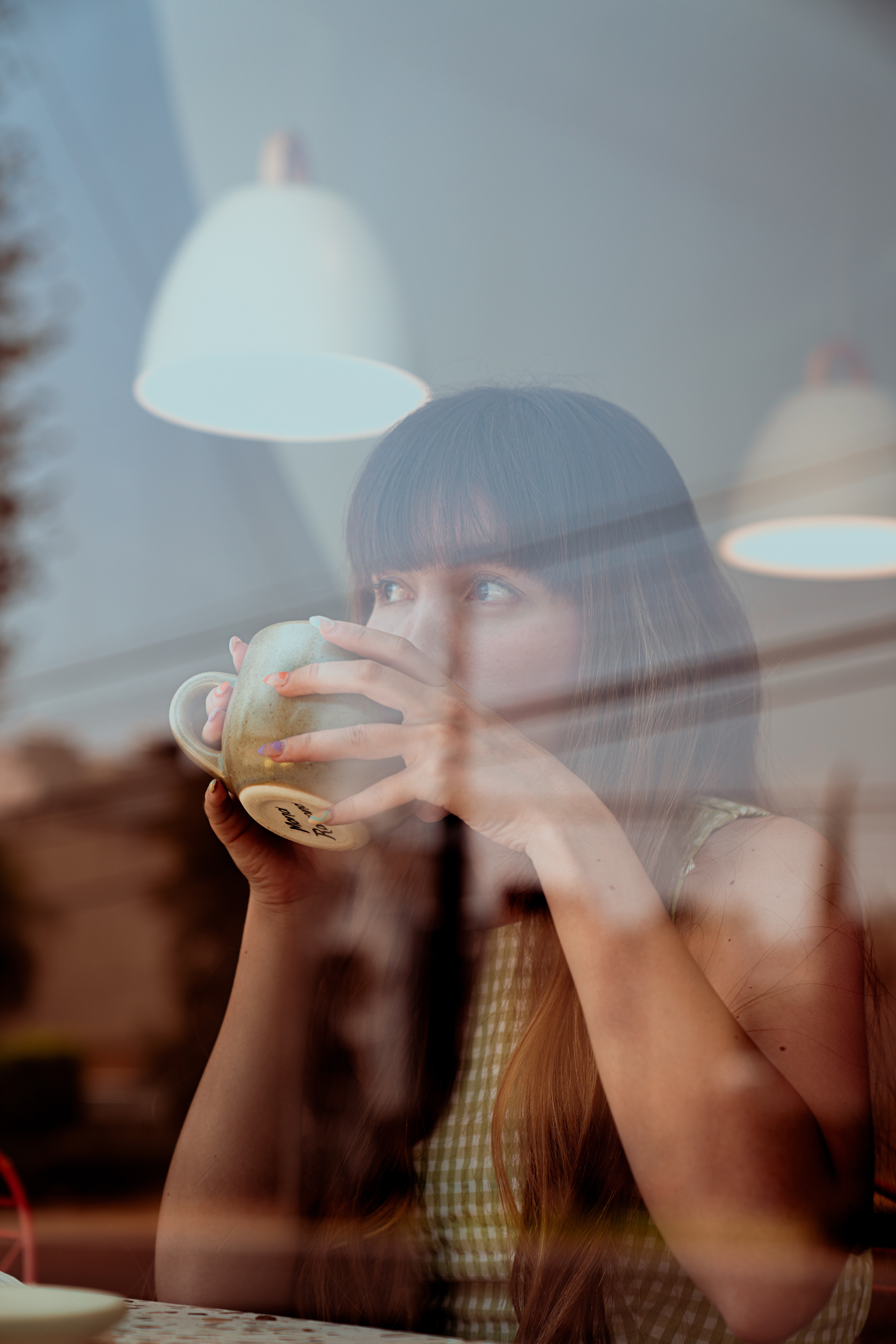 A woman with long hair and bangs holding a cup, looking thoughtfully through a window with reflections of outside elements.