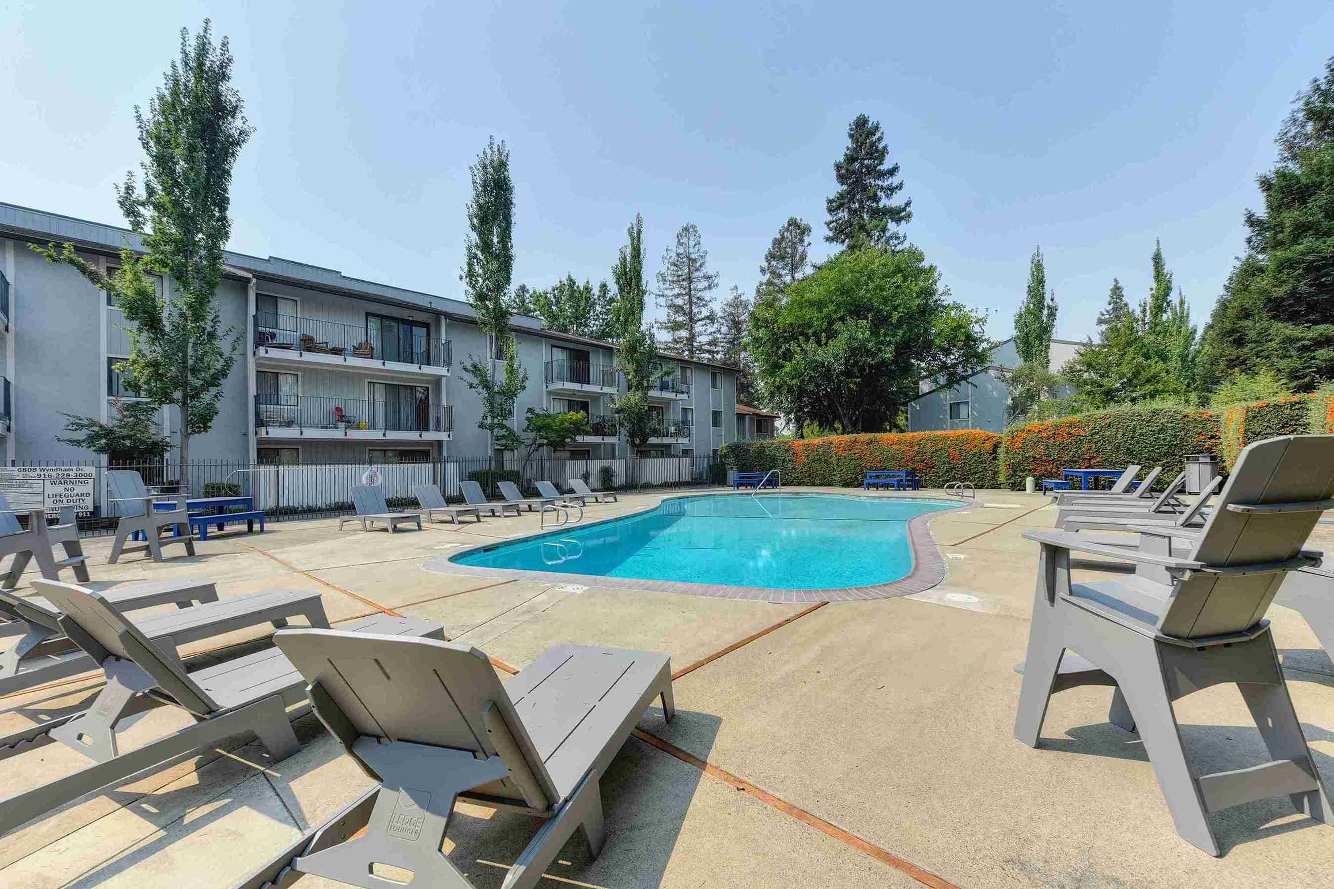 A swimming pool area surrounded by lounge chairs and trees, with low-rise buildings in the background.