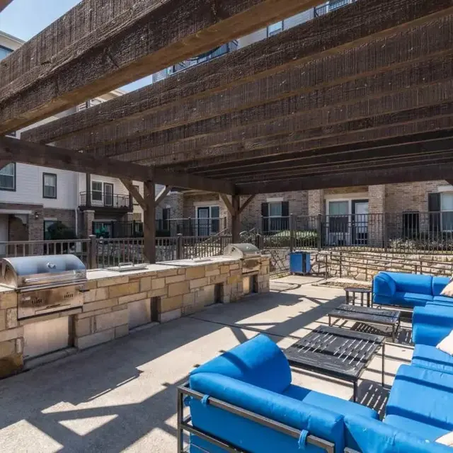 An outdoor lounge area with blue couches and a grill under a wooden pergola, surrounded by apartment buildings.