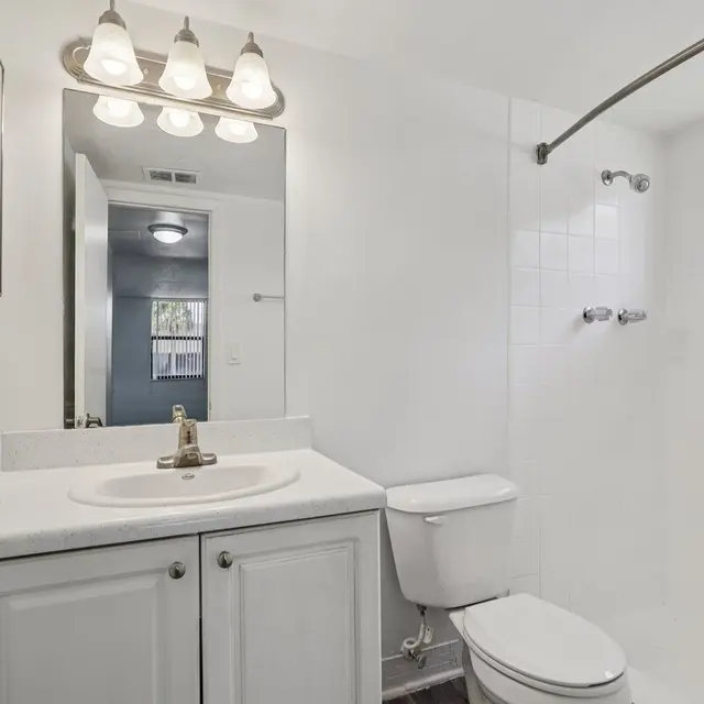 Modern Bathroom Design A clean, modern bathroom featuring a white vanity with a mirror, a sink, a toilet, and a shower area with glass walls. The lighting includes five fixtures above the mirror, providing bright illumination. The walls are painted white, and there's a small window allowing natural light in.