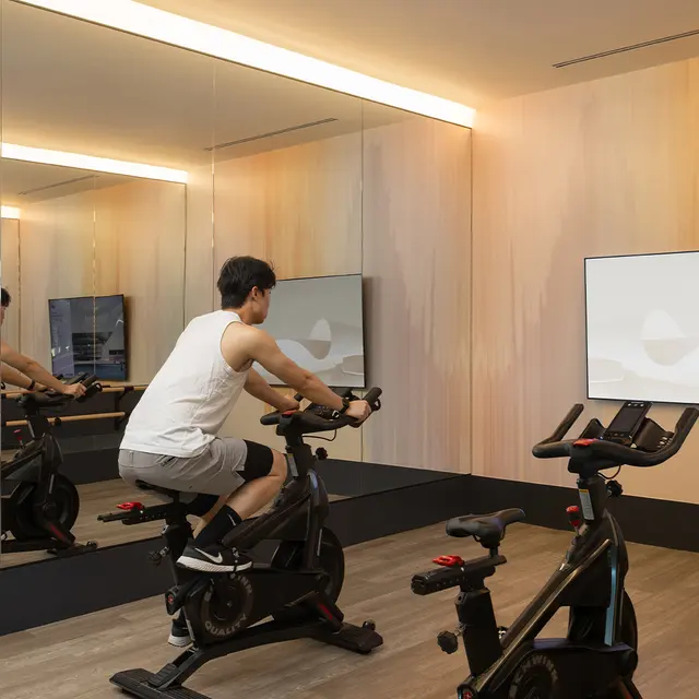 A person exercising on a stationary bike in a modern gym with mirrors and a large TV screen displaying a workout