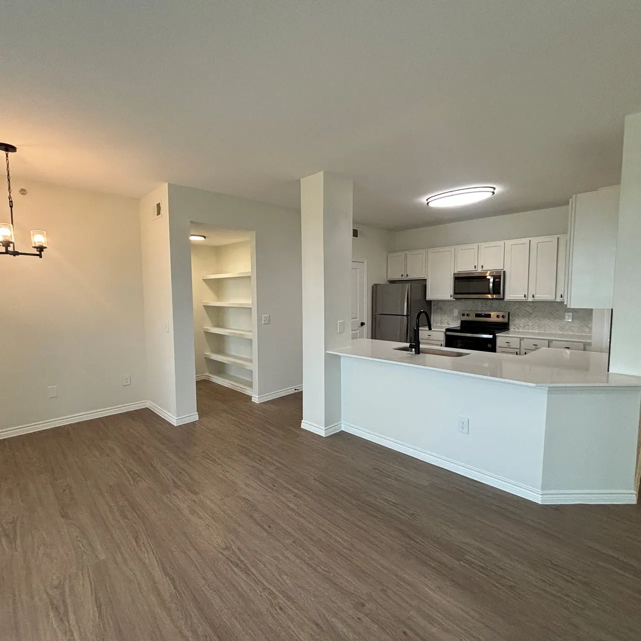 Interior view of a modern apartment with an open layout, featuring a kitchen with white cabinetry and stainless steel appliances, and a living area with wood-like flooring.