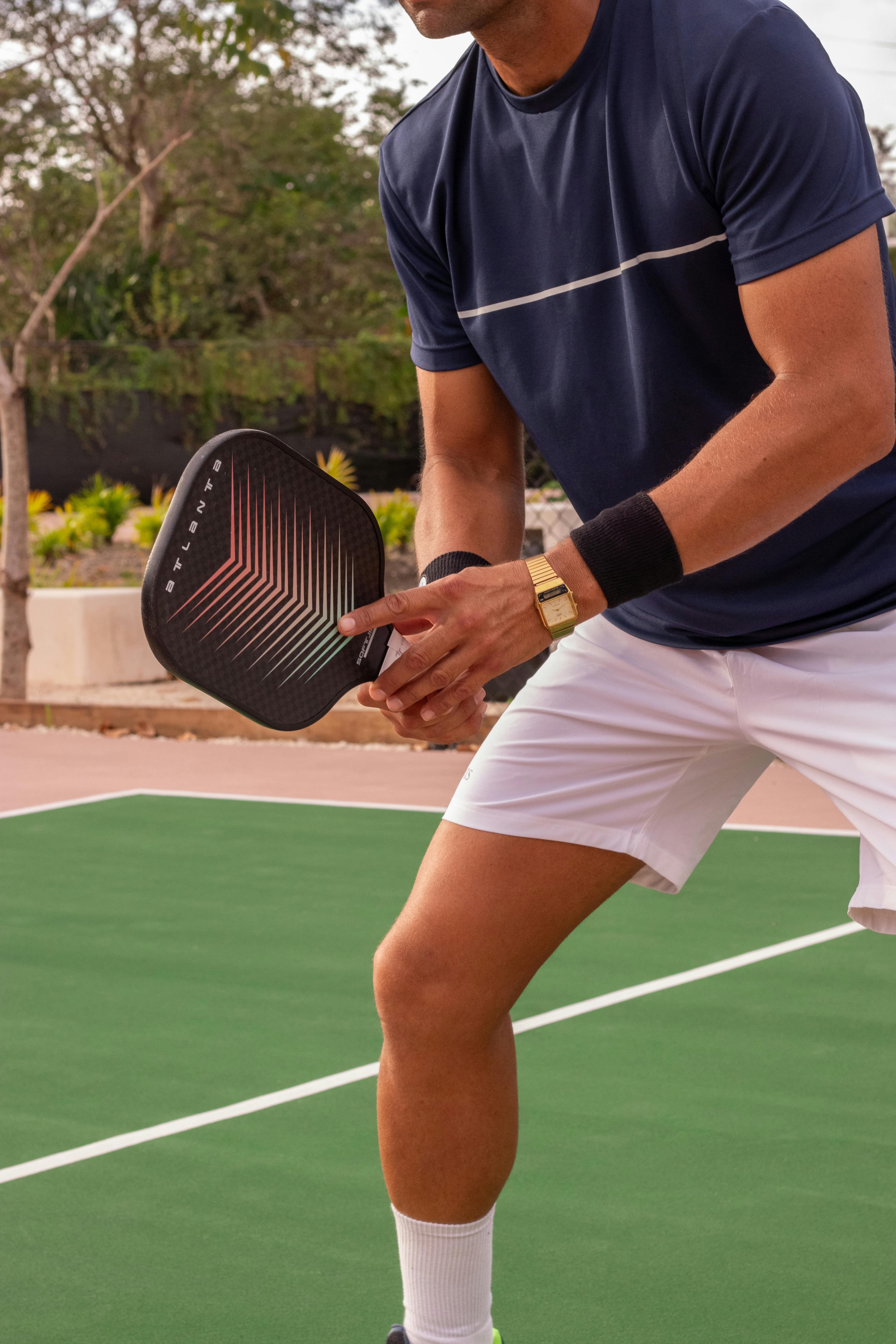 A man holding a paddle tennis racket in a ready position on a green court, wearing a dark t-shirt and white shorts.