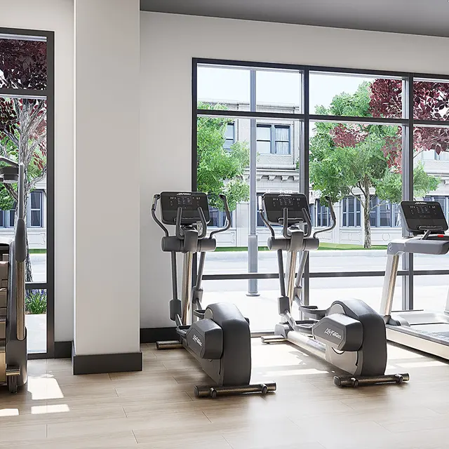A spacious and modern gym interior featuring exercise equipment including treadmills, ellipticals, and stair climbers. Large windows allow natural light to illuminate the space, surrounded by greenery outside.