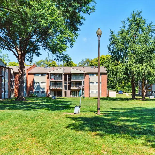 A sunny view of a residential apartment complex with lush grassy areas, large trees, and several two-story brick buildings.