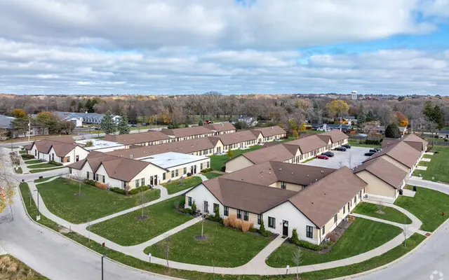 Aerial view of a single-story housing complex with brown roofs and green lawns, surrounded by curved pathways and trees in the background.