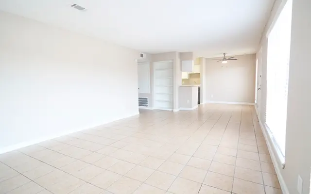 A spacious empty living room with tiled flooring, featuring beige walls and large windows allowing natural light