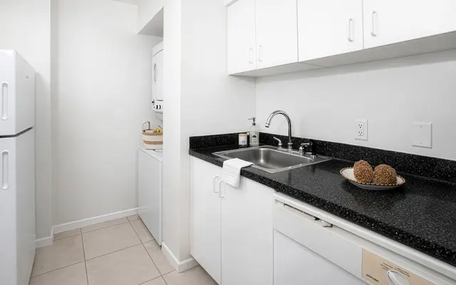 Modern Kitchen Design A modern kitchen featuring a black granite countertop, stainless steel sink, and white cabinetry. On the counter, there is a decorative plate with natural elements. A white refrigerator is visible on the left, and a laundry area is seen in the background.
