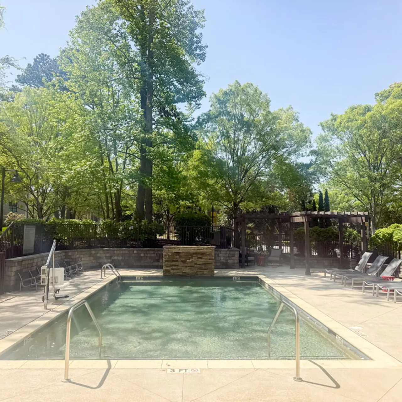 A serene outdoor pool area surrounded by lush green trees, with comfortable lounge chairs nearby and a clear blue sky above.