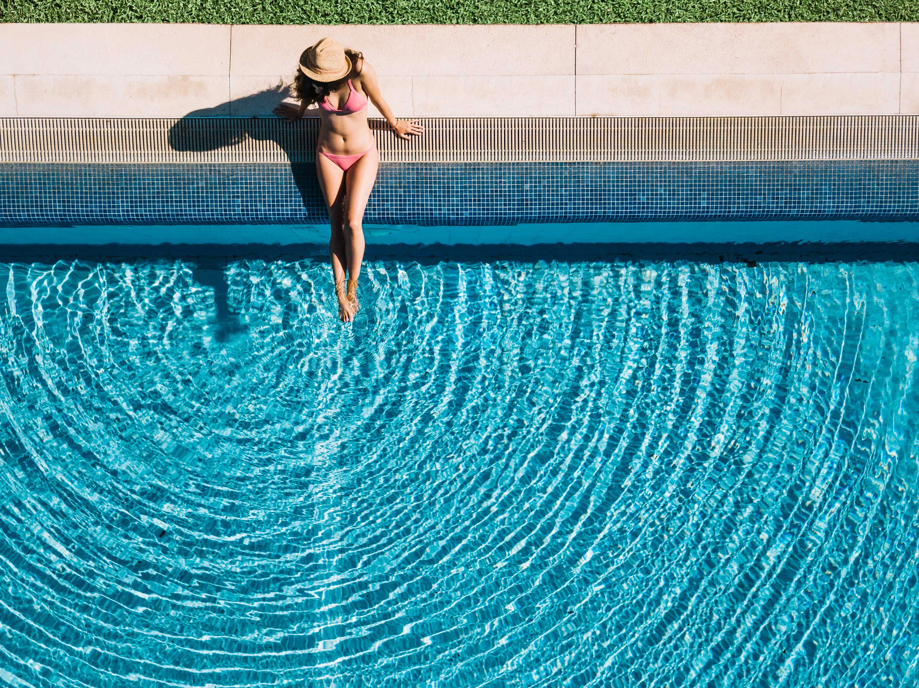 A woman in a pink bikini and wide-brimmed hat relaxing by the edge of a swimming pool, with ripples in the clear blue water.