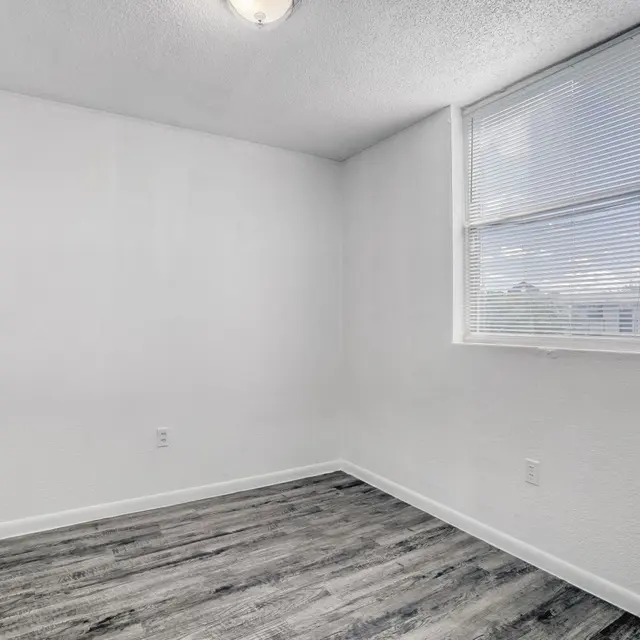An empty room with white walls, a window with blinds, and a wooden floor.