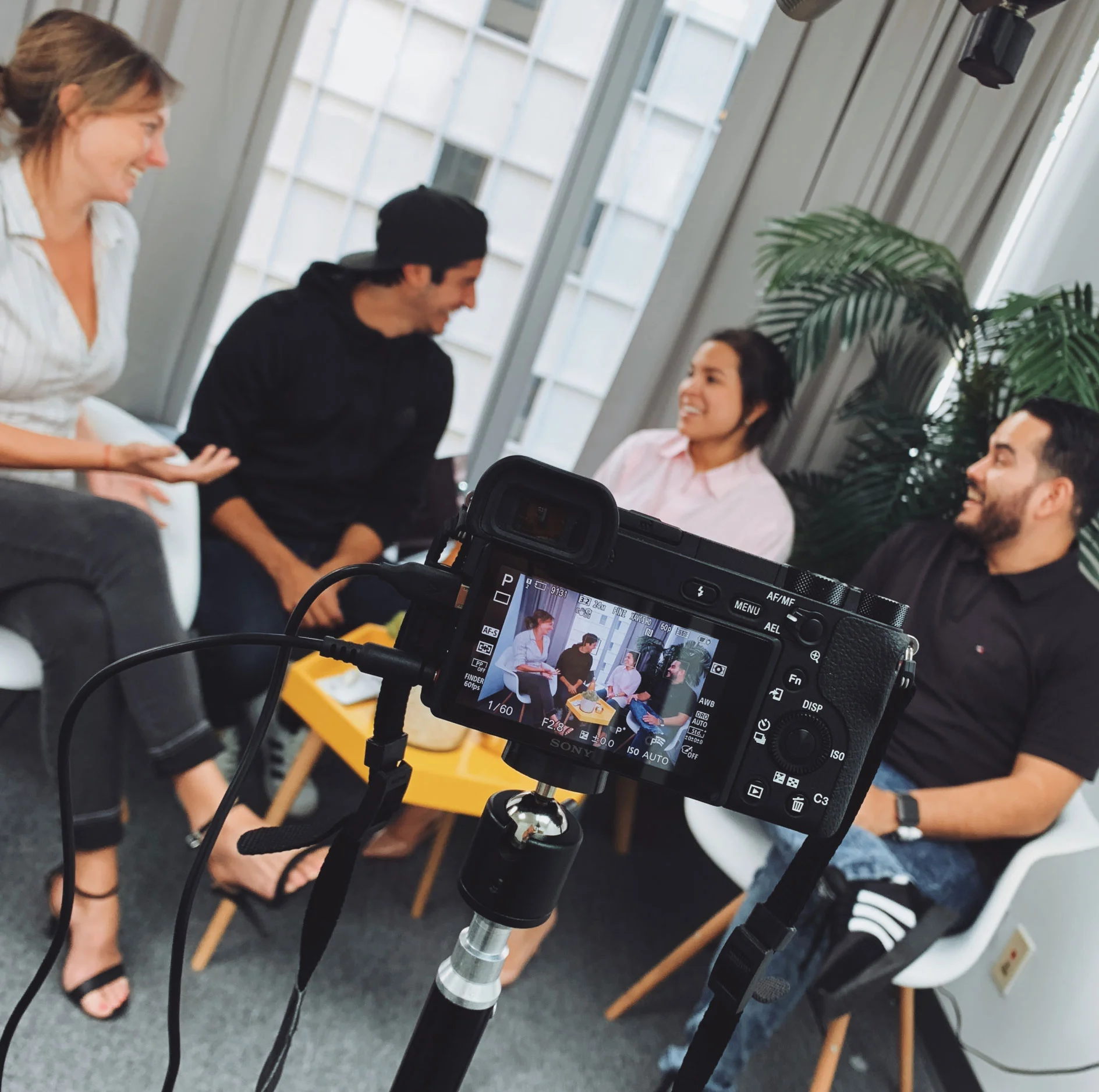 Engaging Group Discussion A group of four people engaged in a discussion, as seen from the perspective of a camera on a tripod capturing the scene. The individuals are sitting in modern chairs with a yellow table in front. They appear to be relaxed and conversing with smiles. The background features large windows and a green plant.