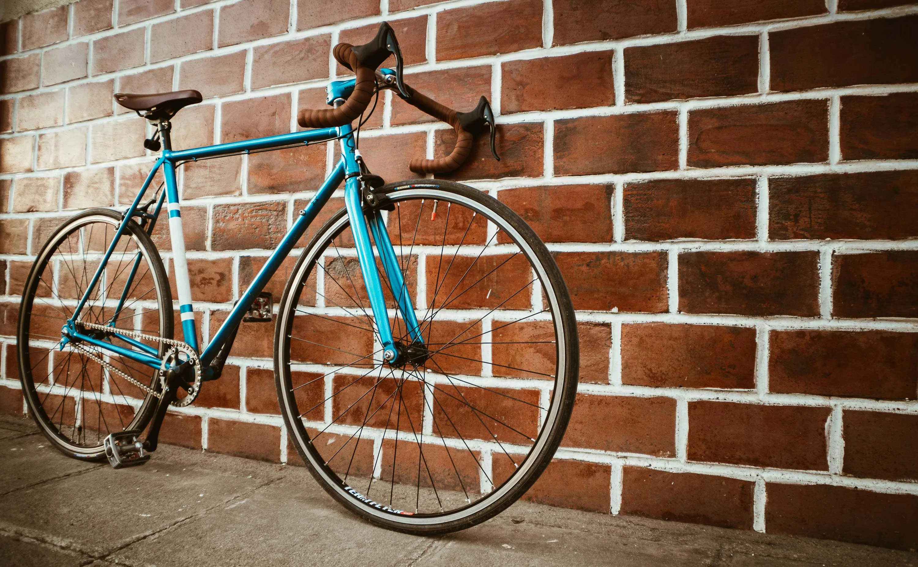 A blue bicycle with brown handlebars leaning against a red brick wall.
