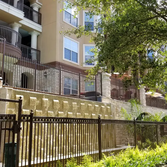 A view of a modern apartment complex with stone walls and a fence, surrounded by greenery.