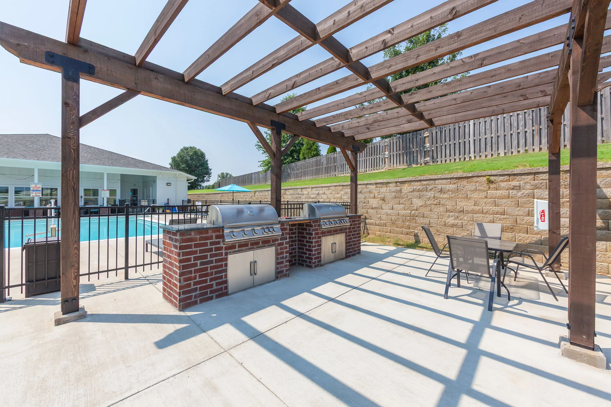 Community Grill Area by Pool An outdoor community area featuring a covered grilling station with two grills, chairs, and a view of a swimming pool in the background.
