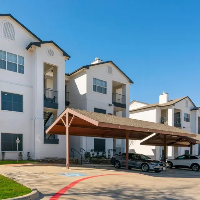 A modern apartment complex with multiple buildings, featuring a white exterior, balconies, and covered parking spaces. Clear blue sky above.