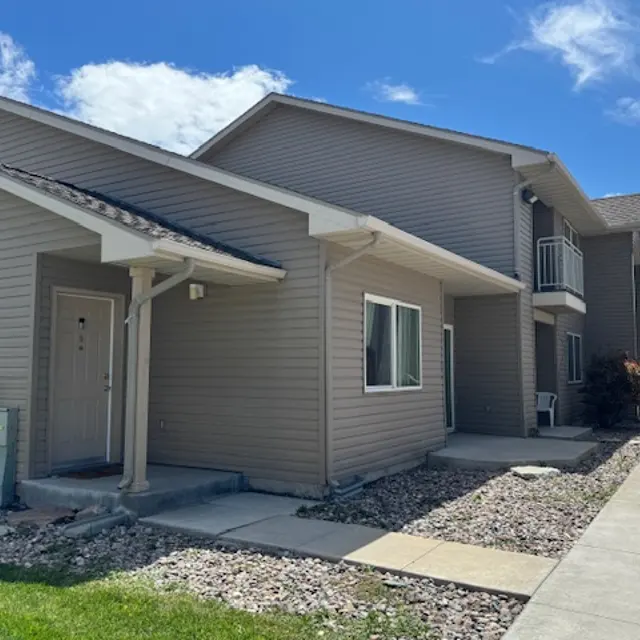 Exterior view of a two-story residential building with a light brown facade and a small porch, surrounded by green grass and a pathway.