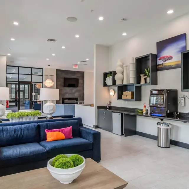 A modern lobby area featuring a navy blue couch with colorful pillows, a coffee table with a white bowl and greenery, stylish wall decor, and a coffee station.