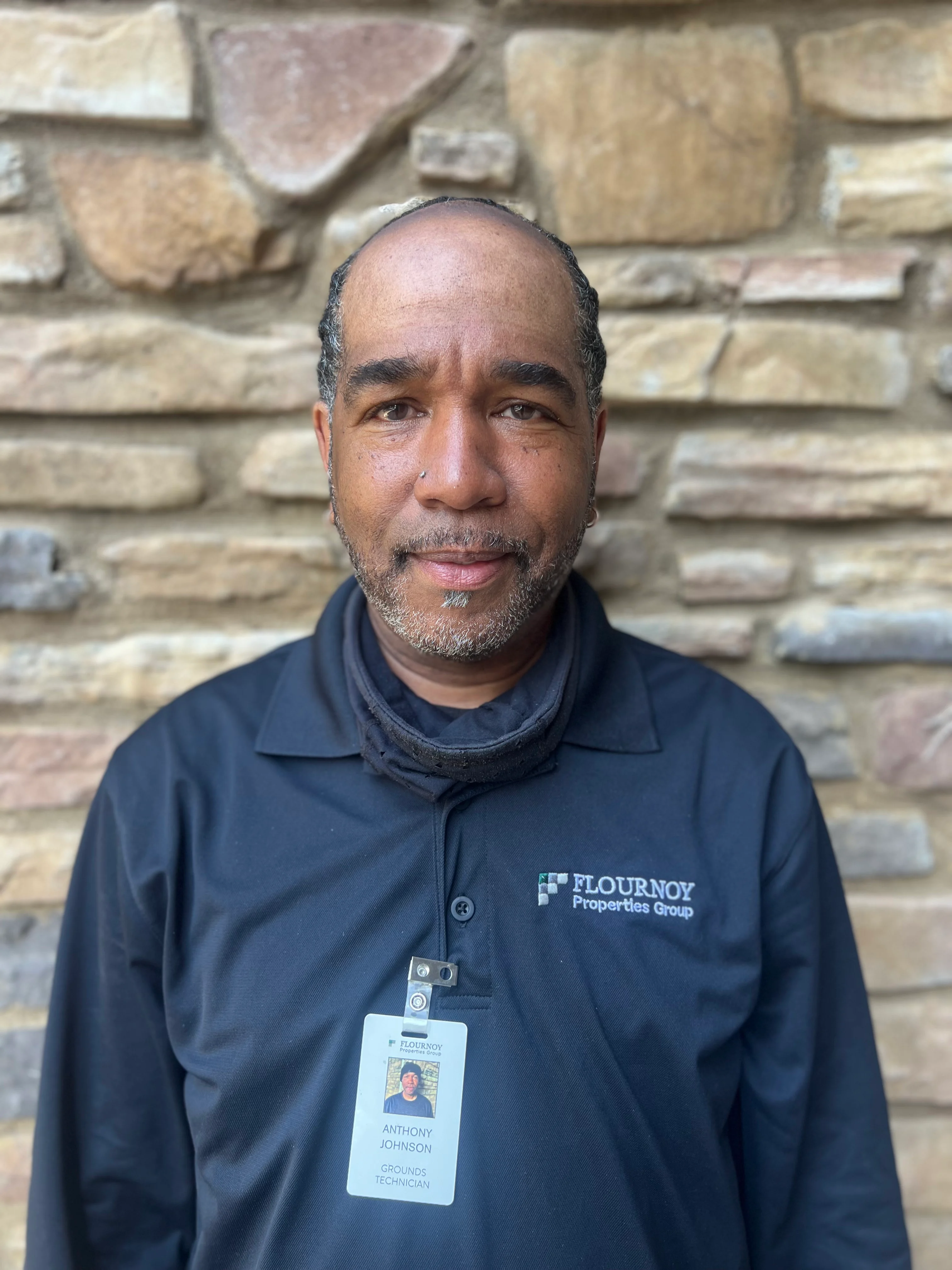 A man with short, slightly curly hair wearing a black collared shirt with a company logo. He has a visible ID badge around his neck and is standing against a stone wall background.