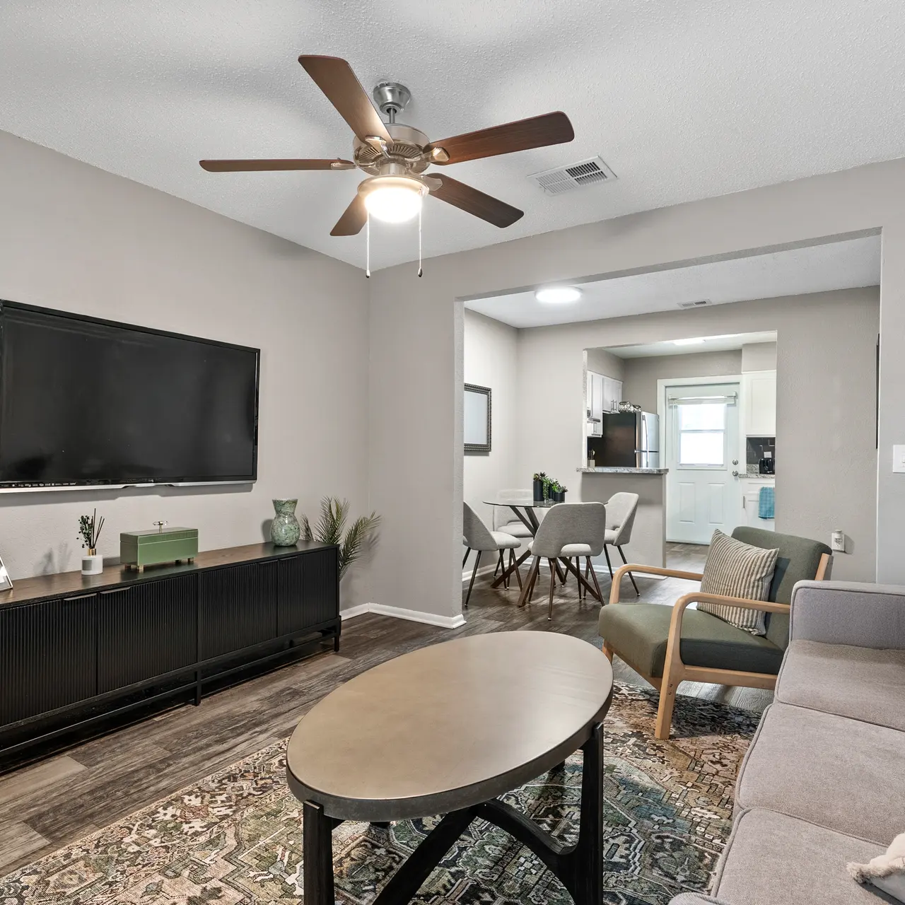 A modern living room featuring a gray sofa, a circular coffee table, a ceiling fan, and a wall-mounted TV. There is a small dining area visible in the background.
