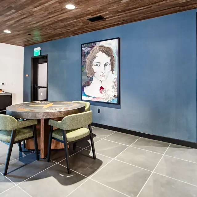 A modern lounge area featuring a circular table with green chairs, a large framed artwork on the wall, and a neat, spacious layout with a tiled floor.