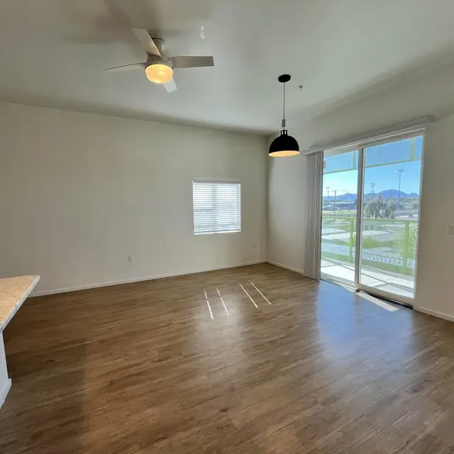 A spacious living room with wooden flooring, a ceiling fan, and large sliding glass doors leading to an outdoor area. A small window is visible on the wall adjacent to the doors.