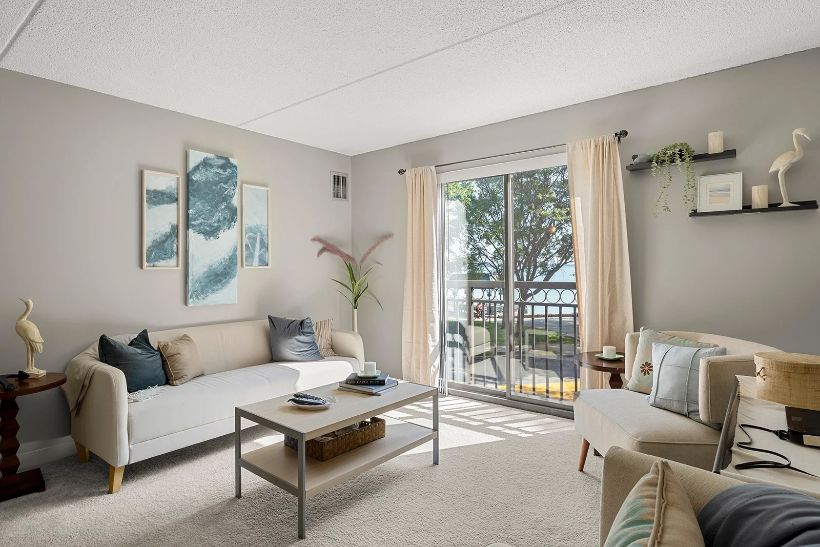 Modern Living Room with Nature View A modern living room featuring a white sofa, decorative pillows, and artwork on the wall, overlooking a balcony with trees outside.