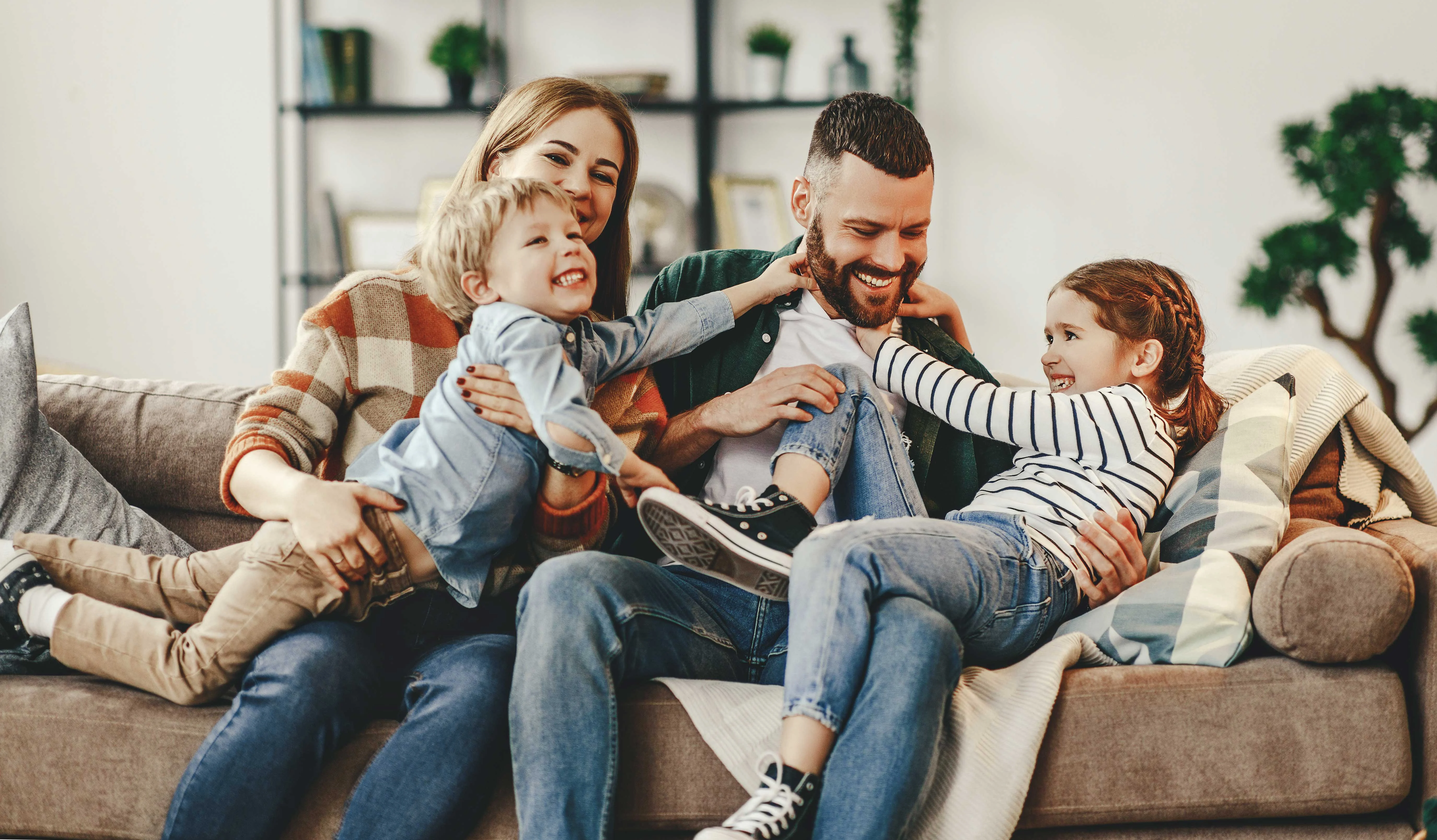 Family Bonding Time A family sitting together on a couch, smiling and playing. A woman holds a young boy on her lap, while a man is playfully interacting with a girl who is sitting next to him. The room has shelves with plants and decor.