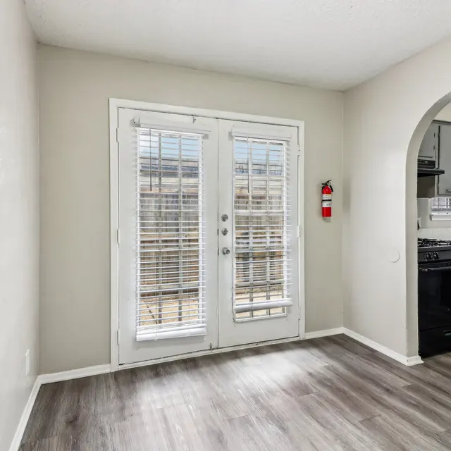 A small dining area with wood laminate flooring, featuring double French doors leading to an outdoor space, and an open view into a kitchen with a black stove and gray cabinets.