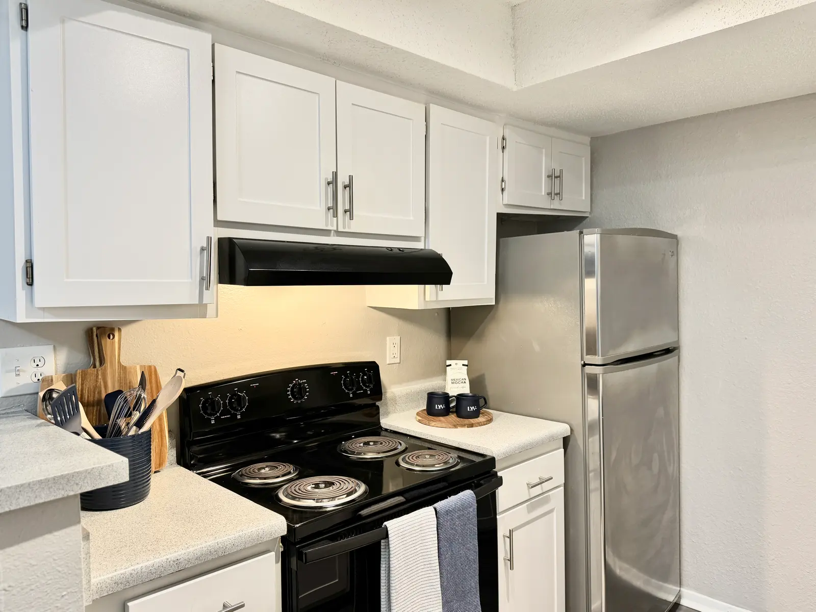 A modern kitchen featuring a black stove, white cabinets, and a stainless steel refrigerator. Kitchen utensils are neatly organized on the counter, with dish towels hanging nearby.